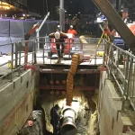 Two construction workers stand on a platform above a deep excavation site at night, supervising another worker inside the pit with large pipes and equipment.