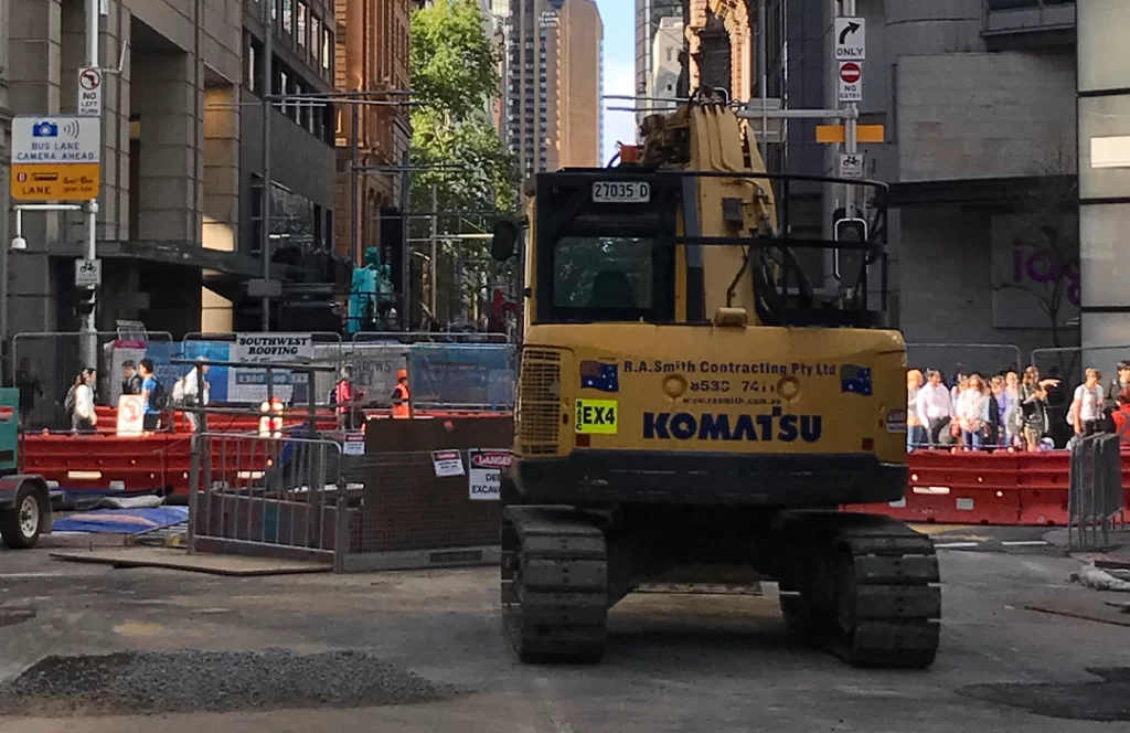 A Komatsu excavator is parked at a city street construction site with barriers, fencing, and people in the background.