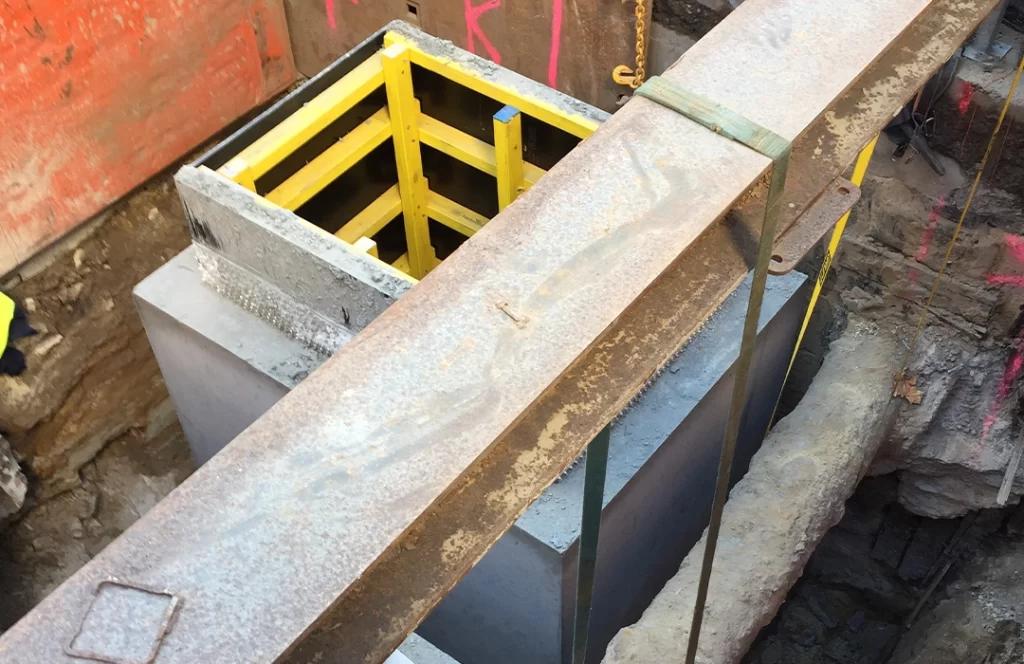 A construction site showing a concrete structure with yellow formwork, surrounded by soil and a large steel beam suspended above.