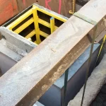 A construction site showing a concrete structure with yellow formwork, surrounded by soil and a large steel beam suspended above.
