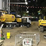 A nighttime city street construction site with excavators, a vacuum truck, safety barriers, and warning signs, illuminated by overhead lights.