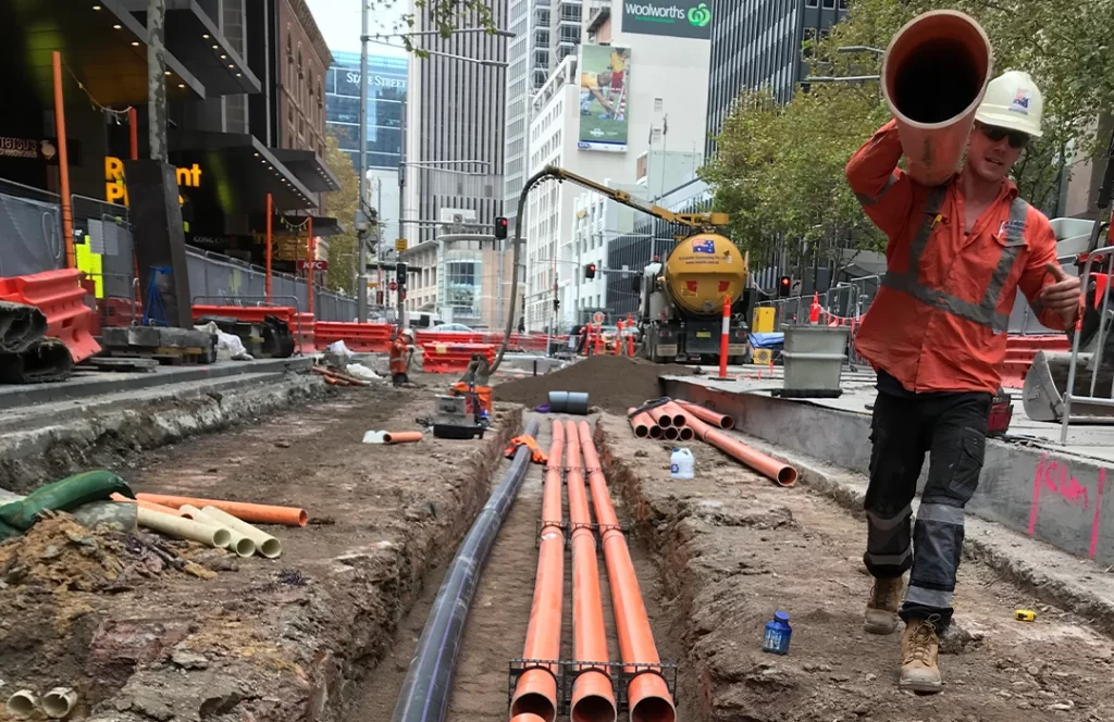 A construction worker in safety gear carries a large orange pipe at a city street work site, with pipes and equipment visible in a dug trench.