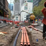 A construction worker in safety gear carries a large orange pipe at a city street work site, with pipes and equipment visible in a dug trench.