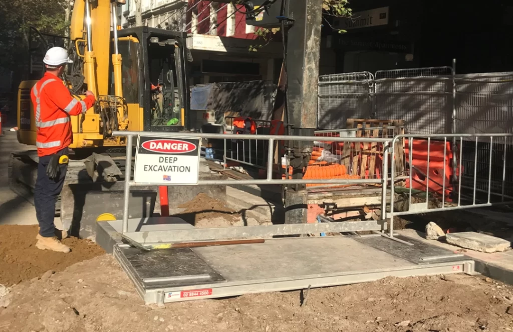 Construction worker stands beside machinery at a fenced-off site with a "Danger Deep Excavation" sign, dirt piles, and equipment visible in sunlight.