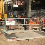 Construction worker stands beside machinery at a fenced-off site with a "Danger Deep Excavation" sign, dirt piles, and equipment visible in sunlight.