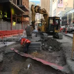 Construction workers in safety gear operate machinery and work in a dug-up urban street, with buildings and signage visible in the background.