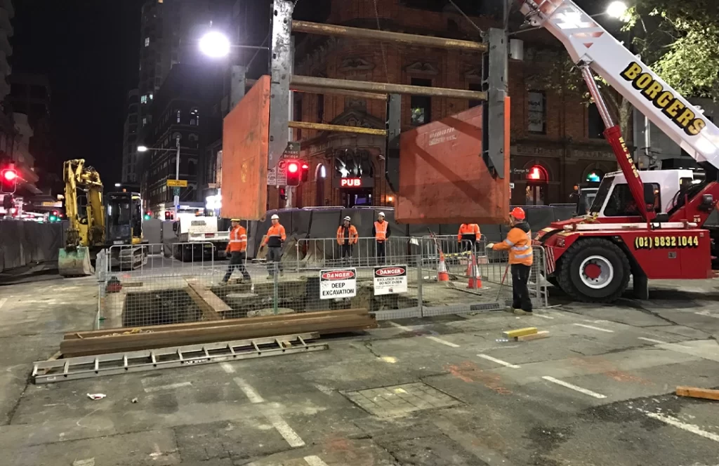 Construction workers in orange vests and helmets work at a fenced-off urban construction site at night, with heavy machinery and warning signs visible.