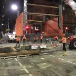Construction workers in orange vests and helmets work at a fenced-off urban construction site at night, with heavy machinery and warning signs visible.