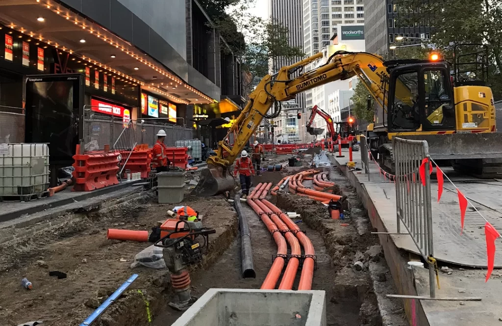 Construction site on a city street with workers, orange pipes being installed underground, excavation equipment, and barriers lining the area.