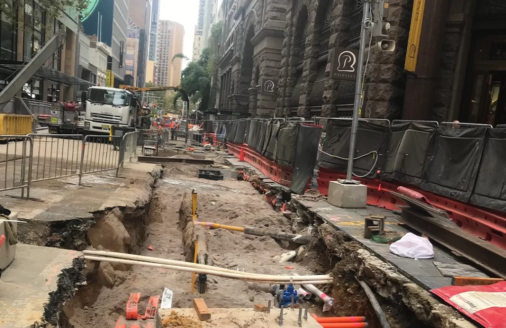 A city street is under construction with a deep trench, exposed pipes, construction barriers, and equipment, surrounded by buildings and partially blocked sidewalks.