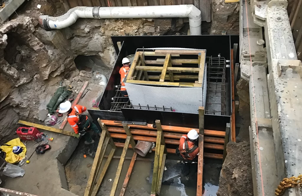 Three construction workers in safety gear work inside an excavated site with reinforced concrete forms, exposed pipes, and construction materials.