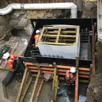 Three construction workers in safety gear work inside an excavated site with reinforced concrete forms, exposed pipes, and construction materials.