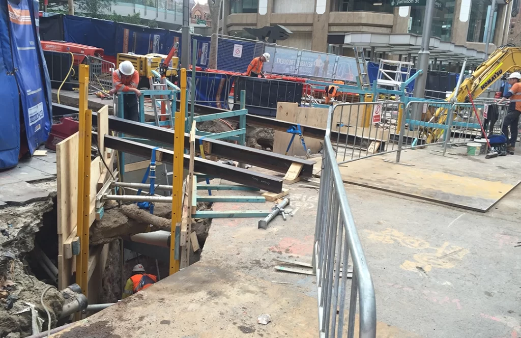 Construction site with workers installing support beams around a large hole in the ground; safety barriers and machinery are present.