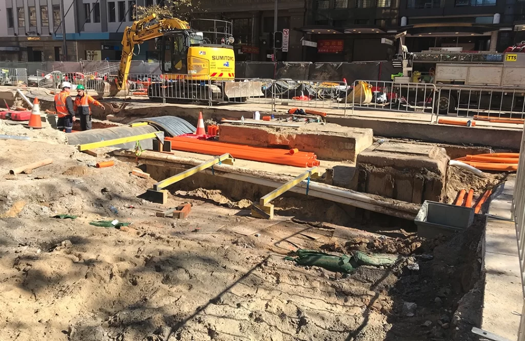 Construction site with exposed trenches, pipes, and cables; workers in safety gear and an excavator operating in the background.