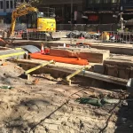 Construction site with exposed trenches, pipes, and cables; workers in safety gear and an excavator operating in the background.