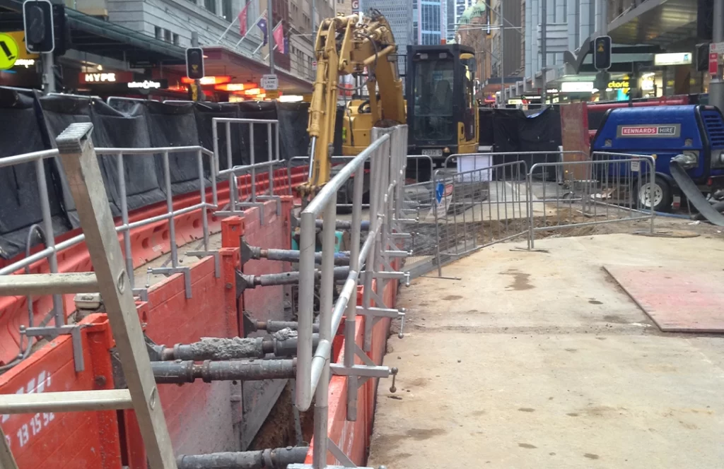 City street construction site with barriers, fencing, and heavy machinery, including a backhoe, blocking part of the sidewalk and road.