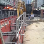City street construction site with barriers, fencing, and heavy machinery, including a backhoe, blocking part of the sidewalk and road.