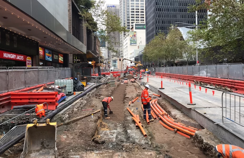 Construction workers install orange utility pipes in a city street excavation zone, surrounded by safety barriers and tall buildings.