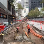 Construction workers install orange utility pipes in a city street excavation zone, surrounded by safety barriers and tall buildings.